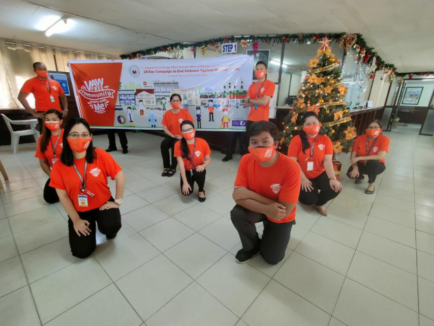 2 Consular personnel pose behind the 18 day campaign official banner and Christmas tree adorned with orange colored ornaments